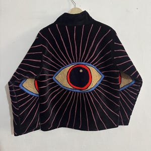Op de afbeelding: Zwarte fluwelen jas met geborduurde oogontwerpen. De jas heeft grote, gestileerde ogen op de rug en mouwen, met rode, blauwe en bruine accenten. Roze lijnen stralen uit de ogen en creëren een sunburst-effect. De jas heeft een hoge kraag.