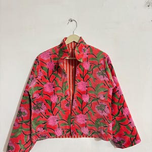 May include: A red floral print jacket with pink flowers and green leaves. The jacket has a high collar and is open in the front, revealing a striped lining. The jacket is hanging on a wooden hanger.