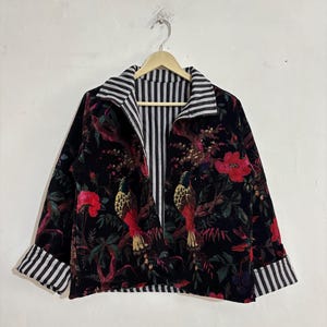 May include: Black velvet jacket with a floral and bird print. The jacket has a striped collar, cuffs, and lining. The design includes red flowers and detailed bird illustrations. The jacket is hanging on a wooden hanger.