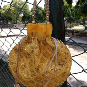 May include: A yellow, round drawstring bag with gold sequins in a diamond pattern. The bag has a yellow fabric top and is suspended by two beaded straps. The drawstring ends in gold tassels. The bag is hanging on a chain-link fence.