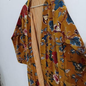 May include: Mustard yellow velvet kimono with a floral pattern in red, blue, and white. The kimono has long sleeves and a striped lining. It is hanging on a wooden hanger.