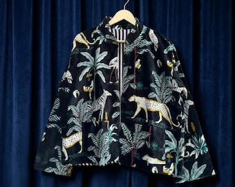 Könnte beinhalten: Schwarze Jacke mit Reißverschluss und lebendigem Dschungel-Print mit Zebras, Leoparden und tropischem Laub. Die Jacke hängt an einem Holzbügel vor einem dunkelblauen Hintergrund. Das Futter der Jacke ist weiß und schwarz gestreift.