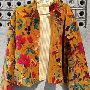 Peut inclure: Veste en velours jaune moutarde avec un motif floral et d'oiseaux rouge, bleu et vert. La veste a une doublure rayée jaune et blanche et est suspendue à un cintre en métal.