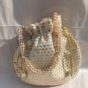 May include: Cream-coloured drawstring bag with gold floral embroidery and a beaded handle. The bag is adorned with small, round gold embellishments and a beaded trim around the edge. A silver-coloured bead is attached to the handle.