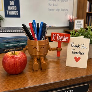 Puede incluir: Un escritorio de madera muestra un arreglo con tem&aacute;tica de profesor. Un portal&aacute;pices sonriente con forma de barril contiene bol&iacute;grafos. Un letrero rojo dice "#1 Teacher" junto a una tarjeta "Thank You Teacher" con un coraz&oacute;n. Una manzana roja y libros completan la escena.