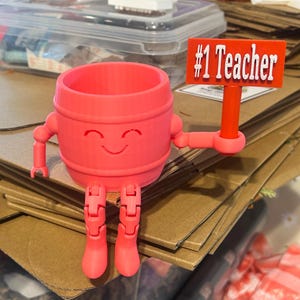 May include: A bright pink 3D-printed barrel-shaped figure with a smiling face, articulated arms, and legs. The figure holds a rectangular red sign with the white text #1 Teacher. The object features a hollow top opening and a smooth finish.