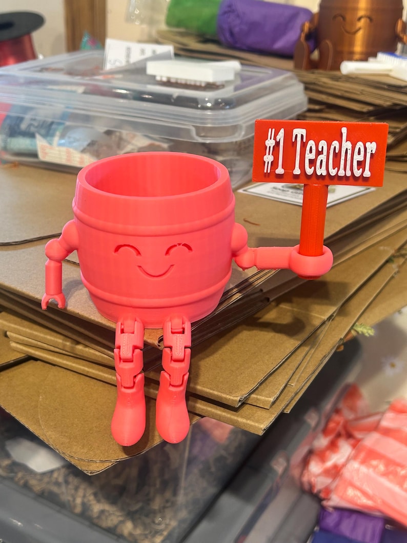 May include: A bright pink 3D-printed barrel-shaped figure with a smiling face, articulated arms, and legs. The figure holds a rectangular red sign with the white text #1 Teacher. The object features a hollow top opening and a smooth finish.
