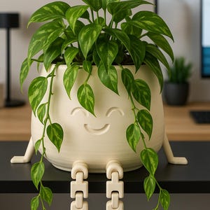 May include: A cream-colored, smiling face planter with attached legs, holding a vibrant green plant with patterned leaves. The plant's vines cascade down, creating a whimsical and decorative indoor plant pot.
