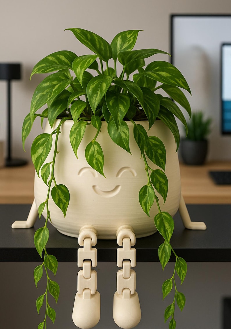 May include: A cream-colored, smiling face planter with attached legs, holding a vibrant green plant with patterned leaves. The plant's vines cascade down, creating a whimsical and decorative indoor plant pot.