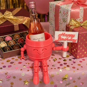 Puede incluir: Un soporte para botella de vino rosa, impreso en 3D, con una cara sonriente, brazos y piernas, que sostiene una botella de rosado. Un letrero que dice "Happy Birthday" se sostiene en su mano. La escena incluye regalos envueltos, bombones y globos.