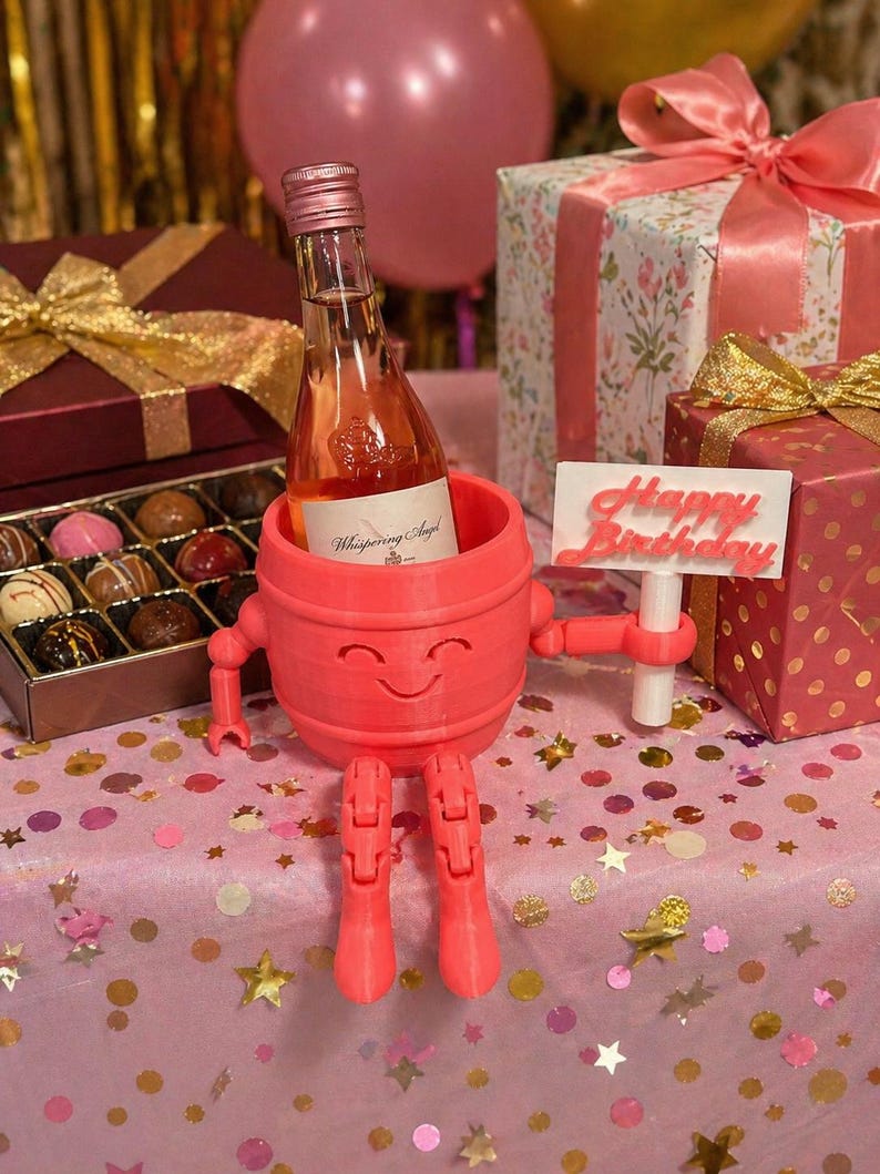 Puede incluir: Un soporte para botella de vino rosa, impreso en 3D, con una cara sonriente, brazos y piernas, que sostiene una botella de rosado. Un letrero que dice "Happy Birthday" se sostiene en su mano. La escena incluye regalos envueltos, bombones y globos.