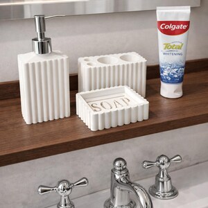 May include: A white bathroom set with a ribbed design, including a soap dispenser, toothbrush holder, and soap dish. A tube of Colgate Total Whitening toothpaste is also present. The set is on a wooden shelf above a chrome faucet.