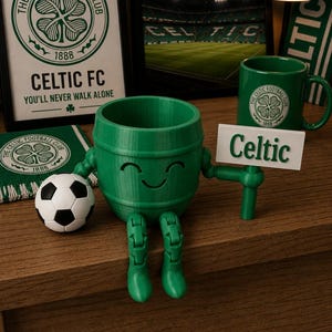 May include: A green 3D-printed Celtic FC barrel-shaped figure with a smiling face, holding a sign that says "Celtic." A soccer ball and a mug with the Celtic logo are also visible. The background includes framed artwork and a scarf with the Celtic FC logo.
