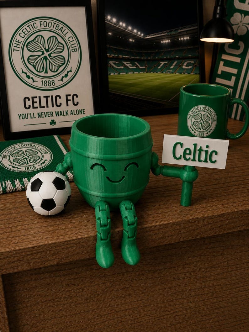 May include: A green 3D-printed Celtic FC barrel-shaped figure with a smiling face, holding a sign that says "Celtic." A soccer ball and a mug with the Celtic logo are also visible. The background includes framed artwork and a scarf with the Celtic FC logo.