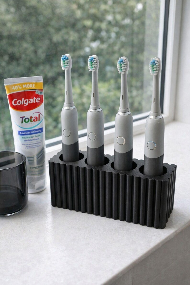 May include: Four electric toothbrushes with gray and black handles are displayed in a black holder. A tube of Colgate Total toothpaste and a dark gray glass are also visible. The toothbrushes have blue and green bristles.