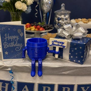 Puede incluir: Una figura azul con forma de barril impresa en 3D con brazos y piernas, sosteniendo un letrero que dice "Happy Birthday". La figura est&aacute; sobre una mesa con una caja de regalo, una tarjeta de cumplea&ntilde;os y una pancarta que dice "Happy Birthday".
