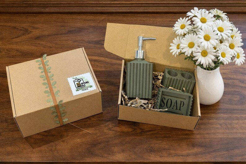 May include: A gift set with a green soap dispenser, soap dish, and soap, presented in a cardboard box with shredded paper. A box with a sticker and a daisy bouquet in a white vase are also present.