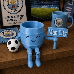 May include: A light blue, 3D-printed figure with a barrel-shaped body, articulated legs, and a smiling face. It holds a sign that reads "Man City." A soccer ball and a mug with the Manchester City logo are also present, along with framed artwork.