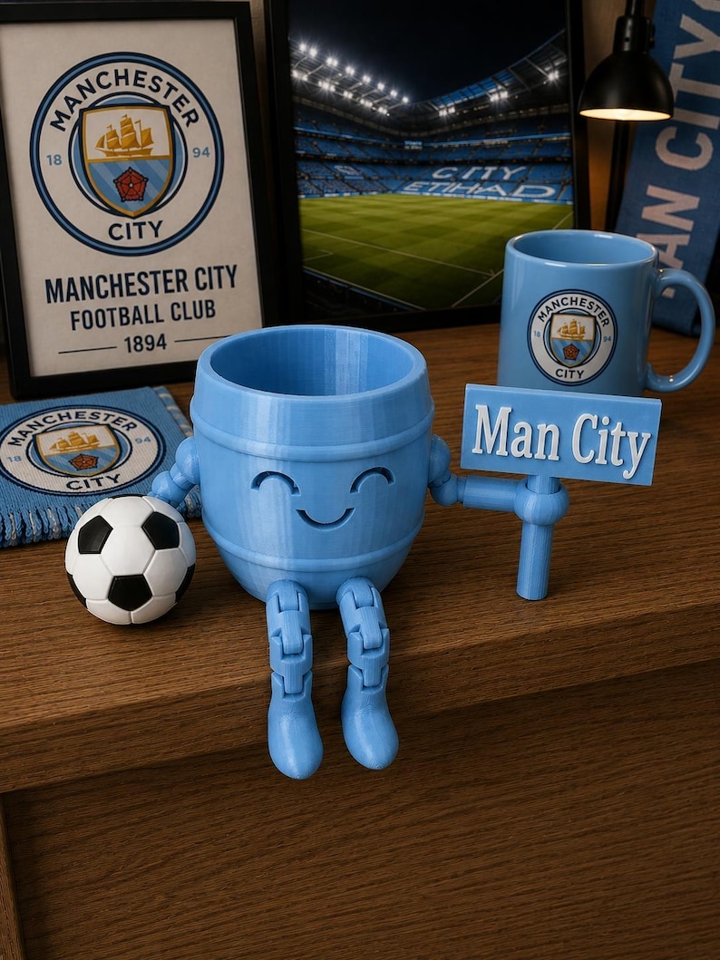 May include: A light blue, 3D-printed figure with a barrel-shaped body, articulated legs, and a smiling face. It holds a sign that reads "Man City." A soccer ball and a mug with the Manchester City logo are also present, along with framed artwork.