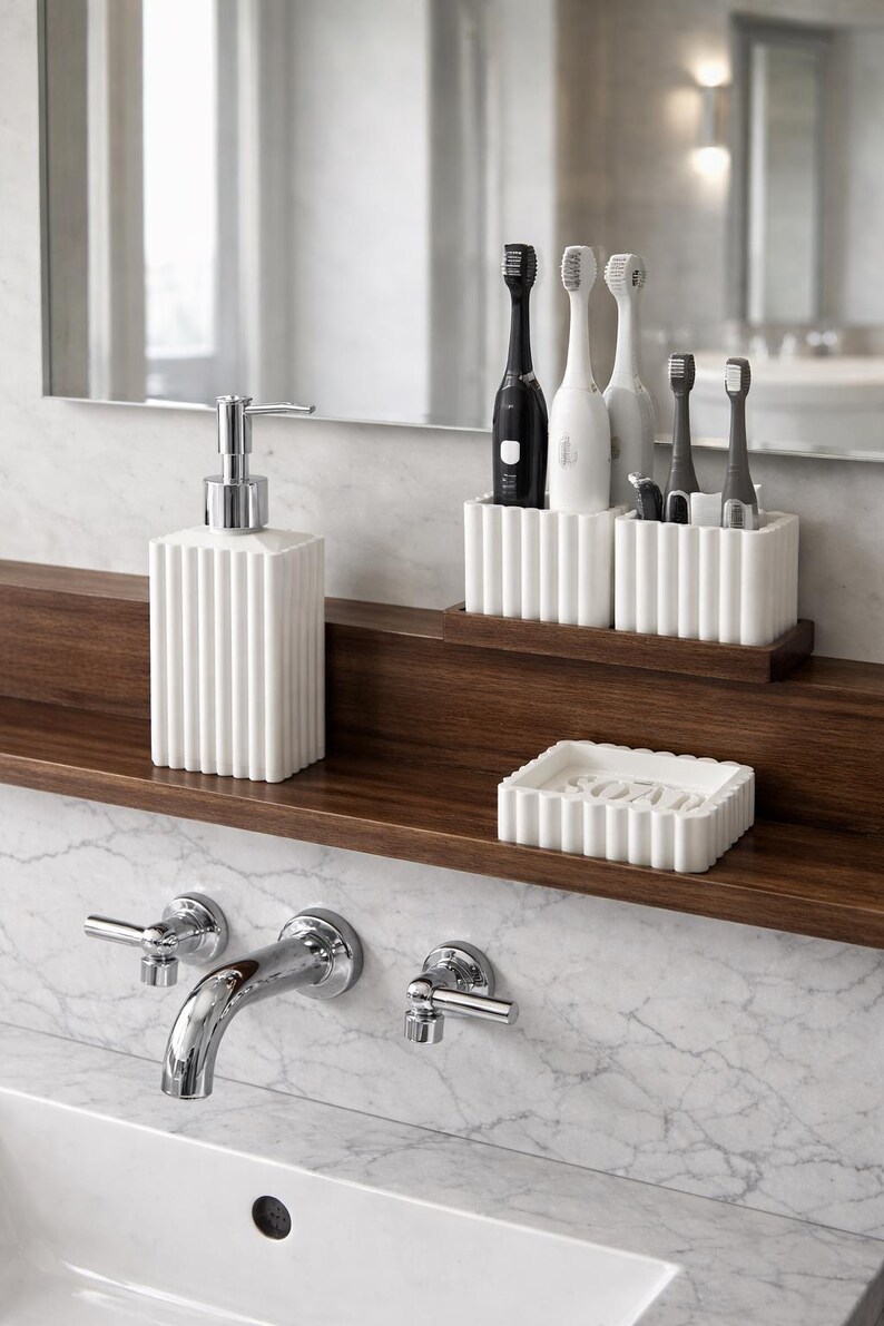 May include: A modern bathroom setup featuring a white soap dispenser, toothbrush holders, and a soap dish. The items are arranged on a dark wooden shelf against a marble wall. The design is minimalist and clean.