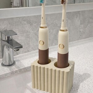 May include: Two electric toothbrushes in a cream and brown color scheme, with a matching holder. The toothbrushes have gold accents and different colored bristles. The holder has a scalloped design and is made of a cream-colored material.