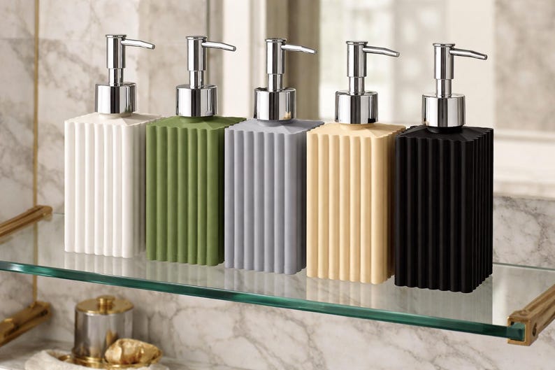 May include: Five rectangular soap dispensers in white, green, gray, beige, and black. Each dispenser has a ribbed design and a silver pump. The dispensers are arranged on a glass shelf.