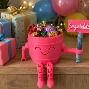 Puede incluir: Un portaregalos rosa en forma de barril con una cara sonriente, brazos y piernas, que sostiene un letrero que dice "Felicidades". El portaregalos est&aacute; lleno de caramelos envueltos y un billete enrollado. Cajas de regalo con lazos en el fondo.