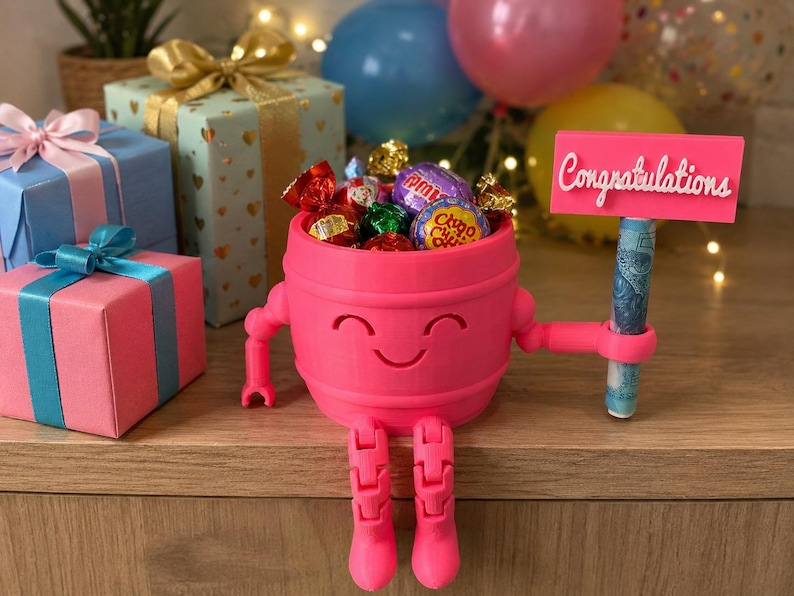Puede incluir: Un portaregalos rosa en forma de barril con una cara sonriente, brazos y piernas, que sostiene un letrero que dice "Felicidades". El portaregalos est&aacute; lleno de caramelos envueltos y un billete enrollado. Cajas de regalo con lazos en el fondo.