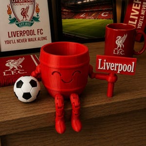 May include: A red, barrel-shaped Liverpool FC themed figurine with a smiling face, boots, and arms holding a sign that says "Liverpool." A soccer ball and a red mug with the Liverpool FC logo are also present. A framed image of the Liverpool FC logo and stadium are in the background.