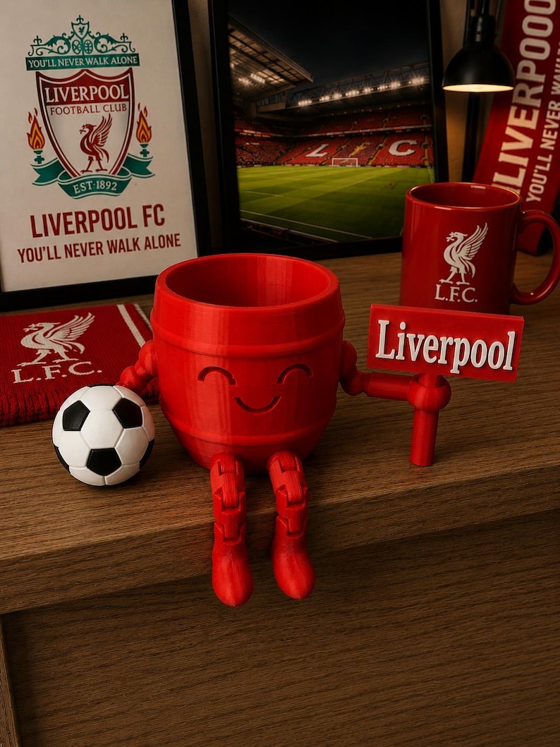 May include: A red, barrel-shaped Liverpool FC themed figurine with a smiling face, boots, and arms holding a sign that says "Liverpool." A soccer ball and a red mug with the Liverpool FC logo are also present. A framed image of the Liverpool FC logo and stadium are in the background.