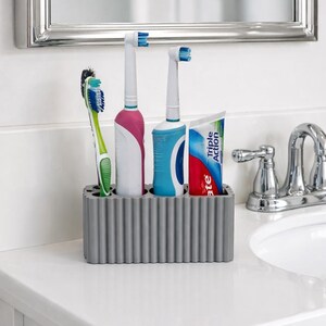 Puede incluir: Un soporte gris para cepillos de dientes con un dise&ntilde;o acanalado contiene un cepillo de dientes manual, dos cepillos de dientes el&eacute;ctricos y un tubo de pasta de dientes. Los cepillos de dientes son blancos, rosas, azules y verdes. El tubo de pasta de dientes es blanco, rojo y azul.