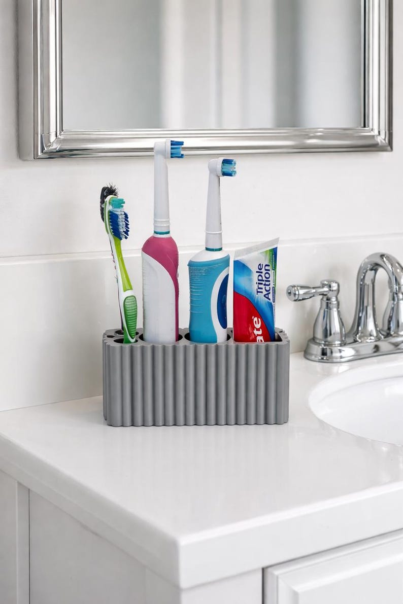 Puede incluir: Un soporte gris para cepillos de dientes con un dise&ntilde;o acanalado contiene un cepillo de dientes manual, dos cepillos de dientes el&eacute;ctricos y un tubo de pasta de dientes. Los cepillos de dientes son blancos, rosas, azules y verdes. El tubo de pasta de dientes es blanco, rojo y azul.