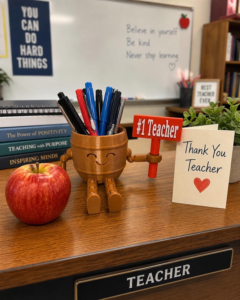 May include: A brown, barrel-shaped pencil holder with a smiling face, articulated arms, and legs. It holds various pens and pencils, and one hand holds a red sign that reads #1 Teacher. Beside it sits a red apple and a card reading Thank You Teacher.