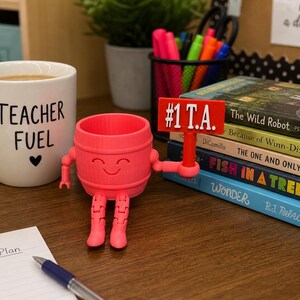 May include: A bright pink, 3D-printed barrel-shaped character with articulated arms and legs, holding a red sign that reads #1 T.A. The character sits next to a white mug labeled Teacher Fuel, a stack of books, and a notepad with a blue pen.