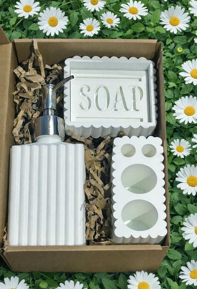 May include: A white bathroom set in a cardboard box, including a ribbed soap dispenser with a silver pump, a soap dish with the word "SOAP", and a toothbrush holder. The set has a scalloped edge design.