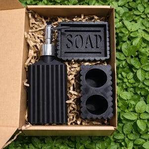 May include: A black bathroom set in a cardboard box, including a soap dispenser, soap dish with the word "SOAP", and a small container. The set is surrounded by brown packing material and set against a green leafy background.