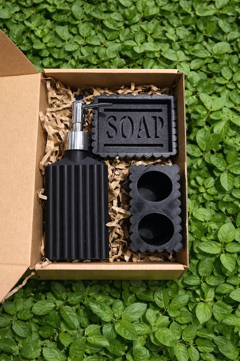 May include: A black bathroom set in a cardboard box, including a soap dispenser, soap dish with the word "SOAP", and a small container. The set is surrounded by brown packing material and set against a green leafy background.