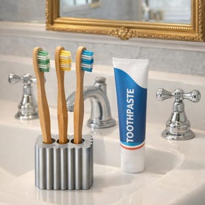 May include: A bathroom sink setup featuring three bamboo toothbrushes with green, yellow, and blue bristles in a silver holder. A tube of toothpaste with the word "TOOTHPASTE" in blue is also present.
