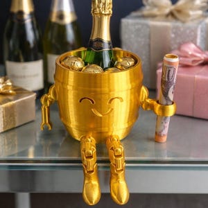 May include: A gold-colored, barrel-shaped champagne bottle holder with a smiling face, arms, and legs. The holder contains a champagne bottle and gold-wrapped chocolates. A rolled-up bill is held in one arm. Champagne bottles and gifts are in the background.