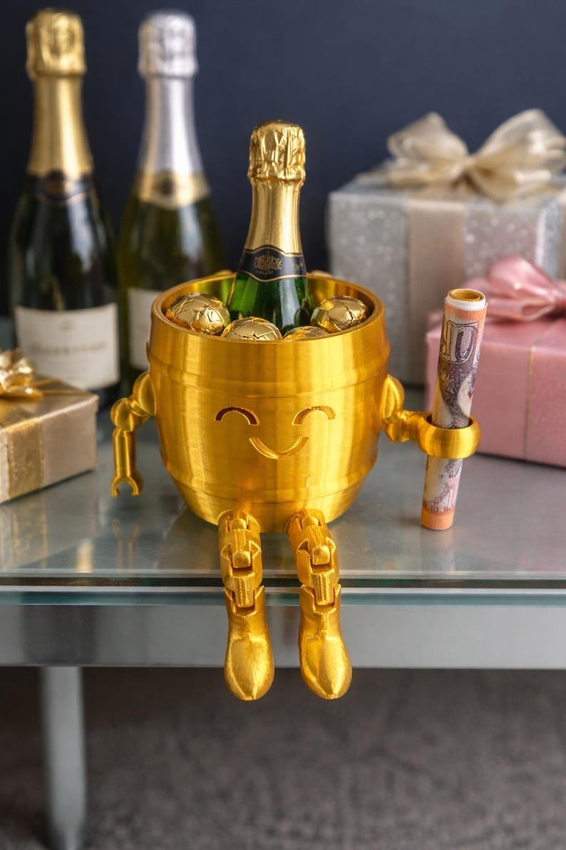 May include: A gold-colored, barrel-shaped champagne bottle holder with a smiling face, arms, and legs. The holder contains a champagne bottle and gold-wrapped chocolates. A rolled-up bill is held in one arm. Champagne bottles and gifts are in the background.