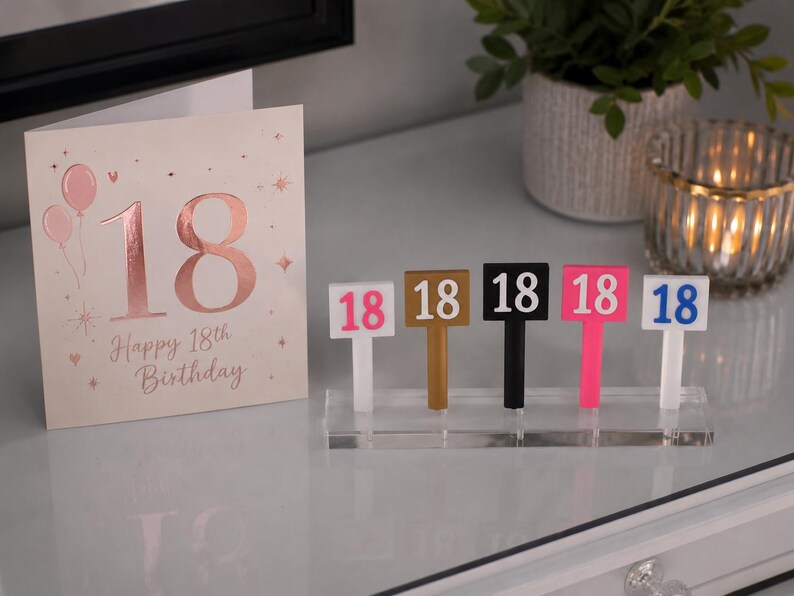 Puede incluir: Tarjeta de cumplea&ntilde;os n&uacute;mero 18 con globos rosas y el texto "Happy 18th Birthday". Junto a ella, cinco peque&ntilde;os letreros con el n&uacute;mero 18 en rosa, dorado, negro, rosa y azul.