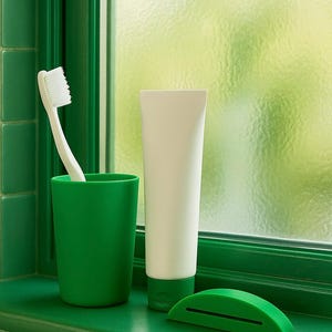 May include: A green toothbrush in a green cup, a white tube of toothpaste, and a green toothpaste tube squeezer sit on a green windowsill. The background is a window with a frosted glass effect.