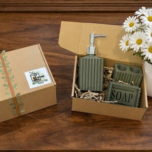 May include: A gift set with a green soap dispenser, soap dish, and soap, presented in a cardboard box with shredded paper. A box with a sticker and a daisy bouquet in a white vase are also present.