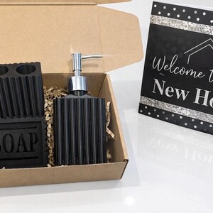 May include: A black bathroom set in a cardboard box, including a soap dispenser, toothbrush holder, and soap dish. The soap dish is labeled "SOAP". A card with the text "Welcome to your New Home" is also included.