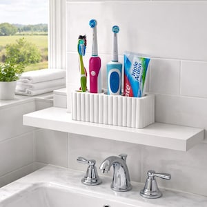 Puede incluir: Un portacepillos de cer&aacute;mica blanca acanalada sobre una estanter&iacute;a blanca, con un cepillo de dientes manual verde y azul, un cepillo de dientes el&eacute;ctrico rosa y azul y un tubo de pasta de dientes. La estanter&iacute;a est&aacute; sobre un lavabo blanco.