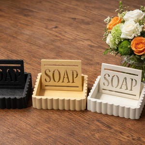 May include: Three decorative soap dishes in black, cream, and white, each with a scalloped edge and a cutout design spelling "SOAP". A bouquet of flowers with orange and white roses sits in the background.