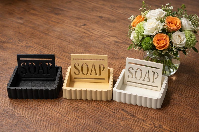 May include: Three decorative soap dishes in black, cream, and white, each with a scalloped edge and a cutout design spelling "SOAP". A bouquet of flowers with orange and white roses sits in the background.