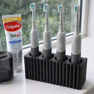 May include: Four electric toothbrushes with gray and black handles are displayed in a black holder. A tube of Colgate Total toothpaste and a dark gray glass are also visible. The toothbrushes have blue and green bristles.