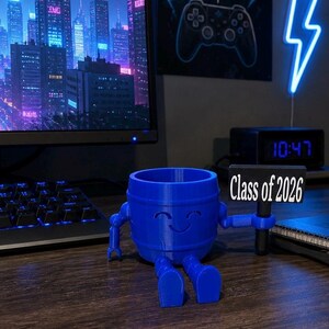 Puede incluir: Un organizador de escritorio azul en forma de barril con brazos y piernas, sosteniendo un letrero que dice "Class of 2026". El organizador tiene una cara sonriente y est&aacute; sentado en un escritorio. Un monitor de computadora y un teclado est&aacute;n en el fondo.