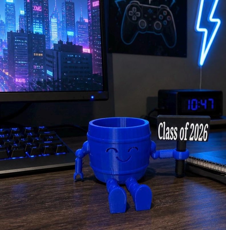 Puede incluir: Un organizador de escritorio azul en forma de barril con brazos y piernas, sosteniendo un letrero que dice "Class of 2026". El organizador tiene una cara sonriente y est&aacute; sentado en un escritorio. Un monitor de computadora y un teclado est&aacute;n en el fondo.